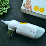 Elektrischer Nasensauger „Beurer“ mit gelbem Einschaltknopf und durchsichtigem Auffangbehälter, daneben ein Karton mit Babyfoto und gelben Symbolen für hygienische Anwendung und einfache Bedienung. Das Gerät liegt auf dunkler Fläche mit Dekoration aus Sukkulenten.