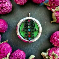 Einzelnes silbernes Duftmedaillon mit Hamsa-Hand-Motiv und bunten Schmucksteinen, auf grünem Filzhintergrund platziert, umgeben von pinken Strohblumen.