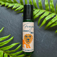 Grüne Glasflasche mit schwarzem Roll-on-Verschluss. Auf dem Etikett steht „Fragantura Löwen Mut – Der Duft, der Mut macht“, begleitet von einer Illustration eines orangefarbenen Löwen mit blauer Nase und freundlichem Gesicht. Die Flasche liegt auf einem dunklen Untergrund, dekoriert mit frischen Farnblättern.