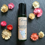 Ein grünes Roll-on-Fläschchen mit schwarzem Deckel liegt mittig auf einem dunklen Untergrund. Auf dem Etikett steht „Fragantura – Mäusezähnchen Zahnungsöl – Sanfte Hilfe für die ersten Zähne“. Eine freundlich wirkende, gezeichnete Maus mit rosa Details ziert die untere Etikettenhälfte. Um das Produkt herum sind bunte Trockenblumen in Rosa, Gelb und Rot arrangiert.