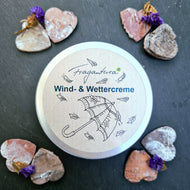 Eine geschlossene Metalldose der Fragantura Wind- & Wettercreme liegt mittig, umrahmt von sechs herzförmigen Holzstücken mit Trockenblumen. Der Deckel zeigt einen Regenschirm mit fallenden Blättern und die Aufschrift „Wind- & Wettercreme“. Der Hintergrund ist aus dunklem Schieferstein.