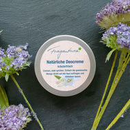 Auf einer dunkelgrauen Steinfläche liegt eine runde Metalldose mit beigefarbenem Etikett. Die Aufschrift auf dem Etikett lautet: „Fragantura – Natürliche Deocreme – kräuterfrisch. Cremen, statt sprühen. Einfach die gewünschte Menge direkt auf die Haut auftragen und einziehen lassen.“ Die Dose ist geschlossen und mittig im Bild platziert. Umgeben ist sie von zarten, violetten Blütenköpfen auf dünnen, grünen Stielen, die dem Arrangement eine natürliche und frische Ausstrahlung verleihen.