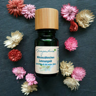 Dunkelgrüne Glasflasche mit Holzdeckel auf dunklem Untergrund, umgeben von rosa, roten und cremefarbenen Trockenblumen. Das Etikett trägt die Aufschrift „Fragantura Mäusezähnchen Zahnungsöl – Sanfte Hilfe für die ersten Zähne“ und zeigt eine zarte Blumenillustration.