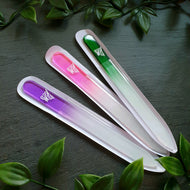 Drei Glasnagelfeilen in den Farben Violett, Rosa und Grün, jeweils mit Farbverlauf zu Weiß, Liliengravur und transparenter Hülle, diagonal angeordnet auf schwarzem Hintergrund mit grüner Blätterdekoration.