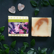 Das Bild zeigt ein rechteckiges Seifenstück der Marke Fragantura neben der dazugehörigen Verpackung. Die Banderole trägt die Aufschrift „Fragantura – Natürliche Duschseife – Blütengarten – Rose Geranie – Lavendel“ und zeigt florale Motive in Rosa und Violett. Die Szene ist mit grünen Blättern und zwei kleinen Herzdekorationen gestaltet.